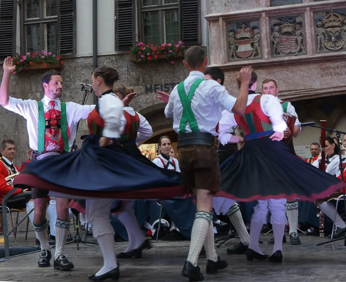 Dancers in Tyrolean costume spin in front of a house front with painted coats of arms in the Old Town of Innsbruck, with musicians seated behind them
