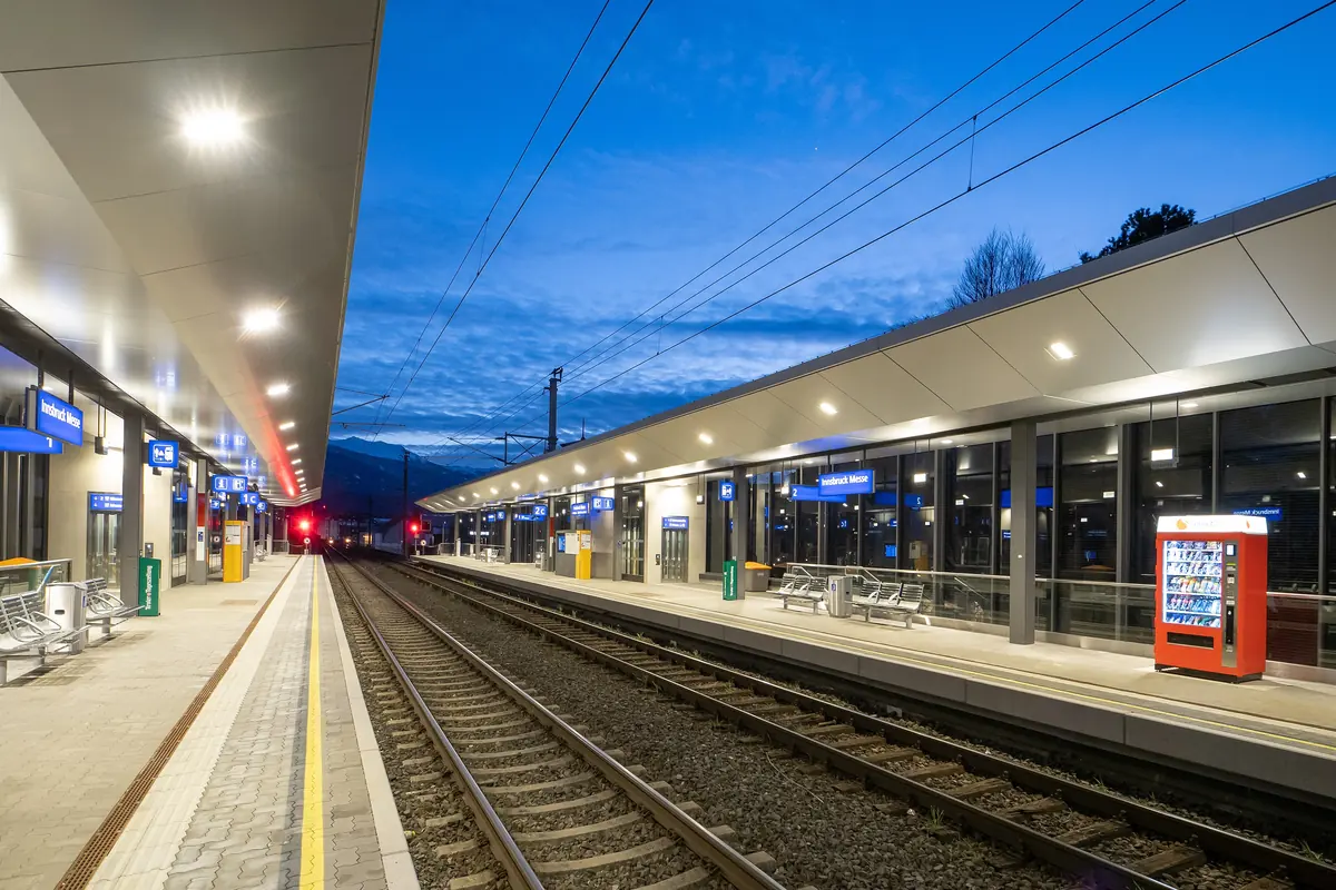 A railway platform at the Innsbruck Messe stop under a deep blue evening sky, with lit canopies along the track