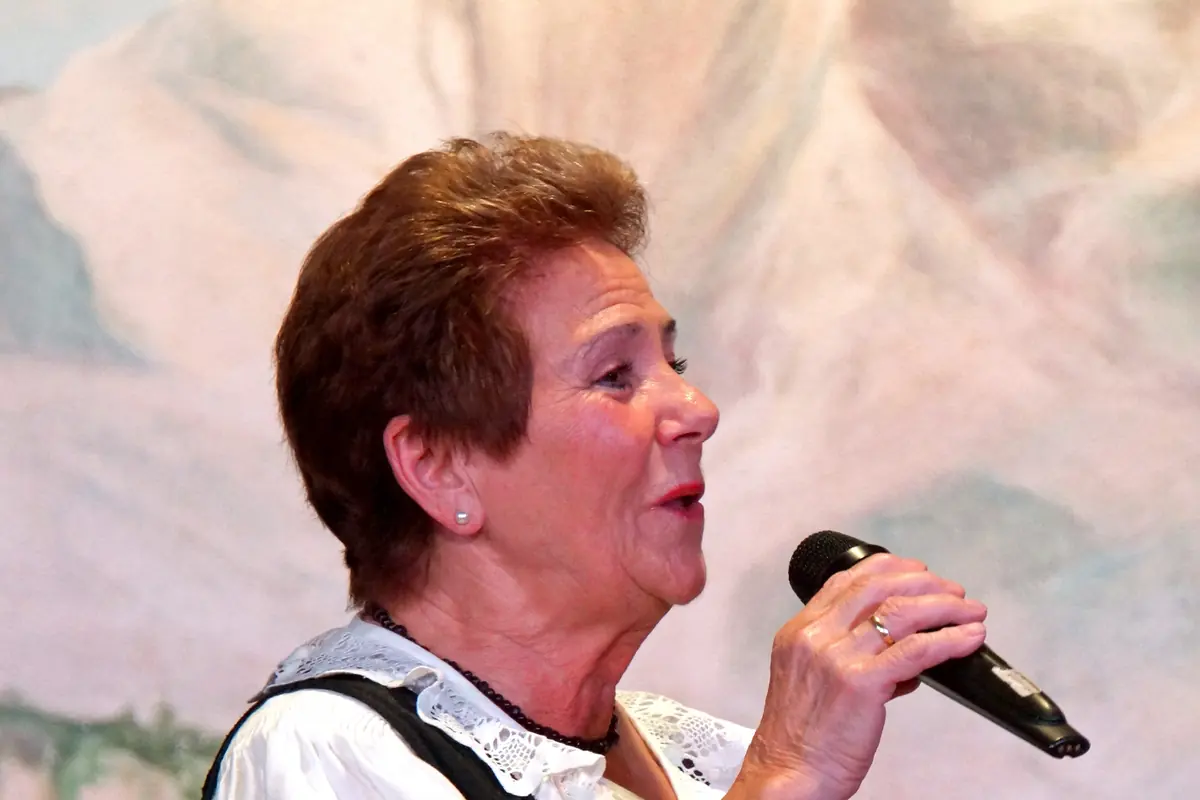 A woman in a dirndl sings into a microphone in front of a painted mountain backdrop